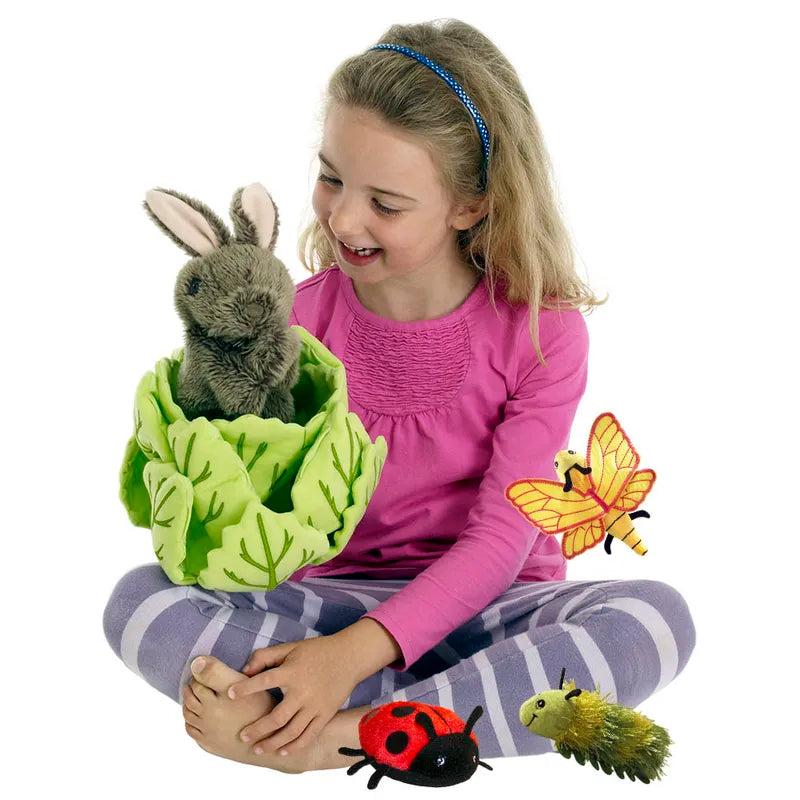 A young blonde girl sitting cross-legged on the floor in a pink top and striped pants smiles at The Puppet Company Hide-Away Rabbit nestled in lettuce, while plush toys of a ladybug, caterpillar, and butterfly add to her whimsical scene.
