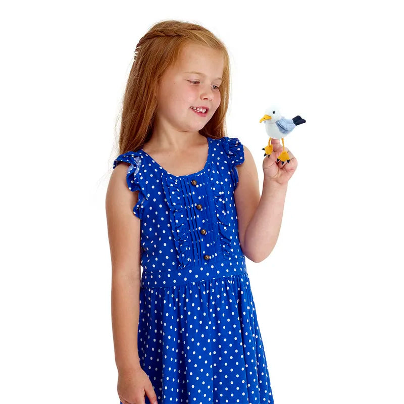 A young girl with long red hair smiles while holding The Puppet Company Seagull Finger Puppet, perfect for storytelling. She is wearing a blue dress with white polka dots and is looking at the soft toy against a plain white background.