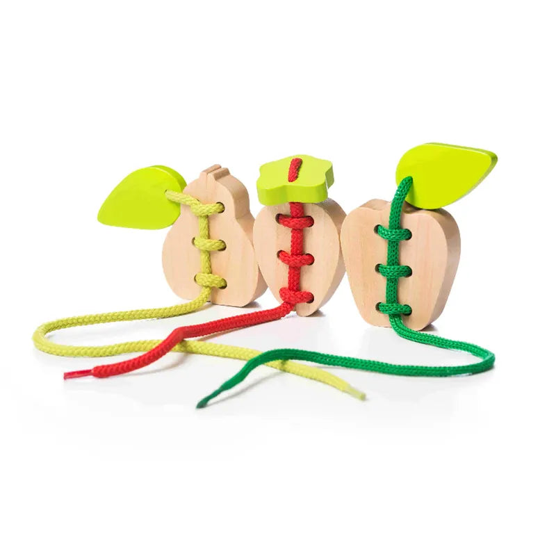 Cubika Lacing Fruits features three chunky wooden fruits with green leaf tops and colored laces (red, yellow, green) that thread through holes—perfect for building fine motor skills and hand-eye coordination.