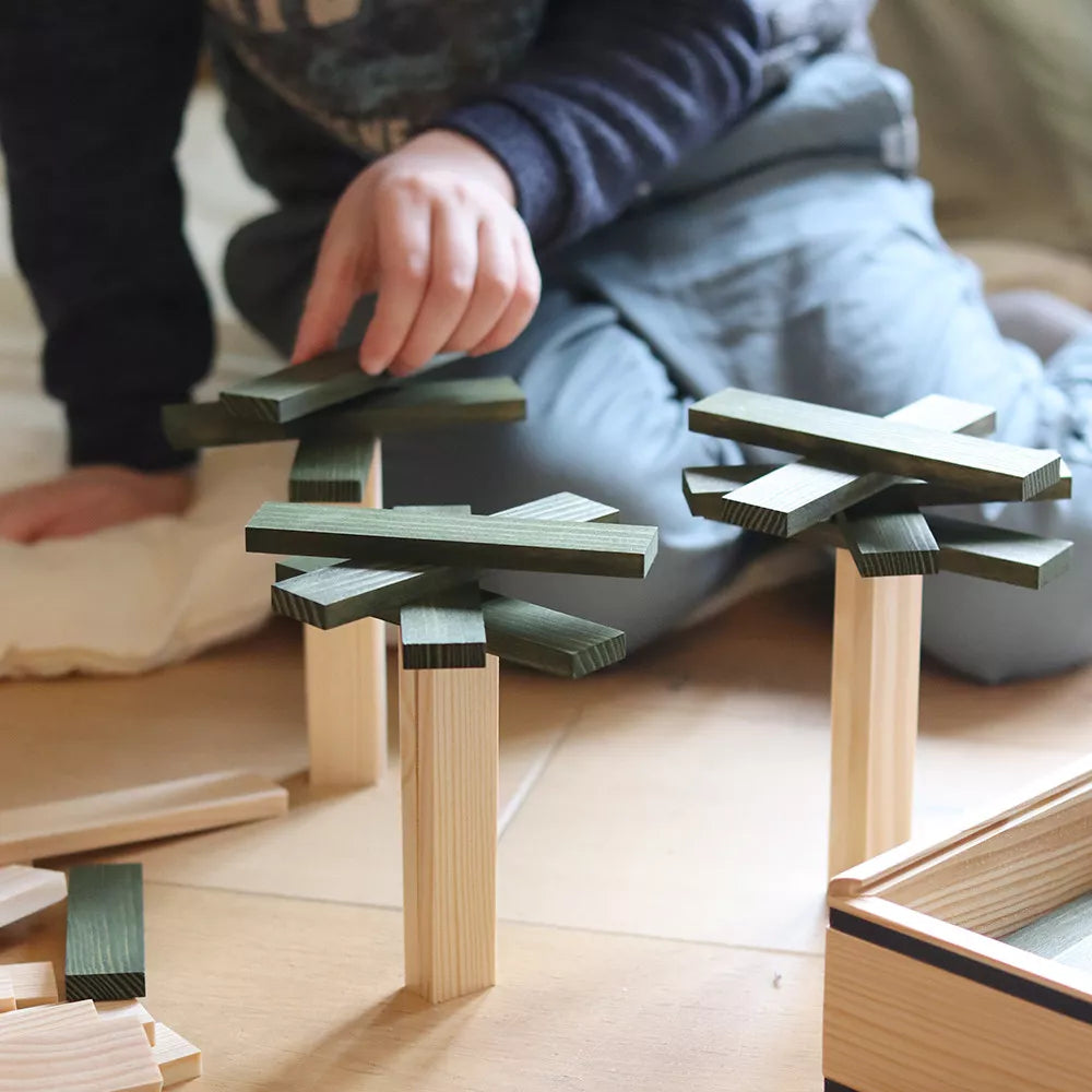 A child sits on the floor, stacking green and natural wooden blocks into tree-like shapes, engaging in creative play with a wooden construction set.