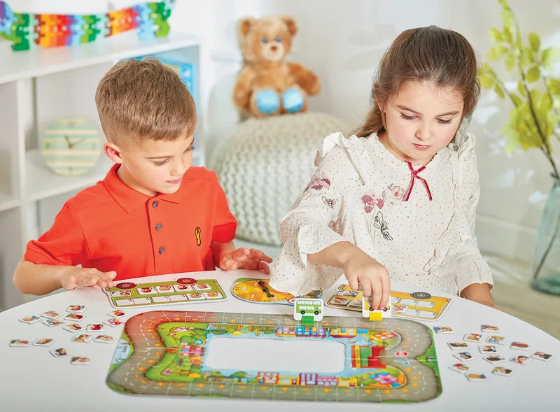 Two children are engaged in a game of Orchard Toys Bus Stop at a table. The boy, dressed in a red shirt, is focusing intently on his pieces, while the girl, wearing a white dress, places a yellow piece onto the board. In the background, a stuffed teddy bear can be seen.