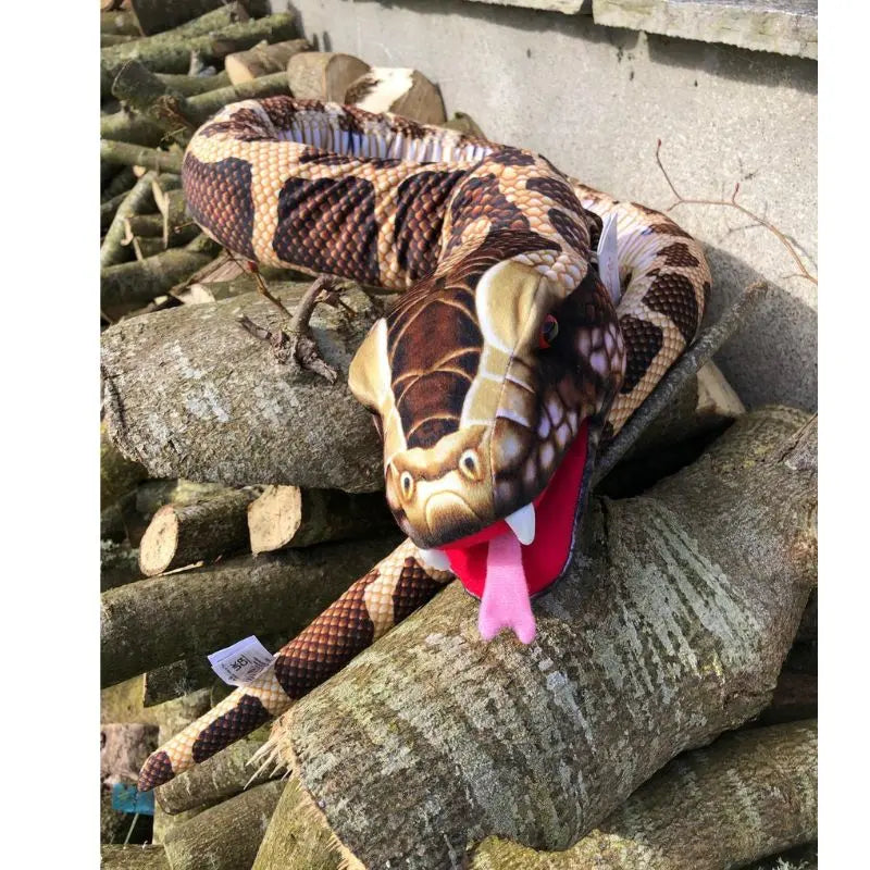 The Puppet Company Large Creature Snake Brown, with its mouth open and pink tongue out, lies on a pile of cut logs against a concrete wall, looking impressively lifelike.