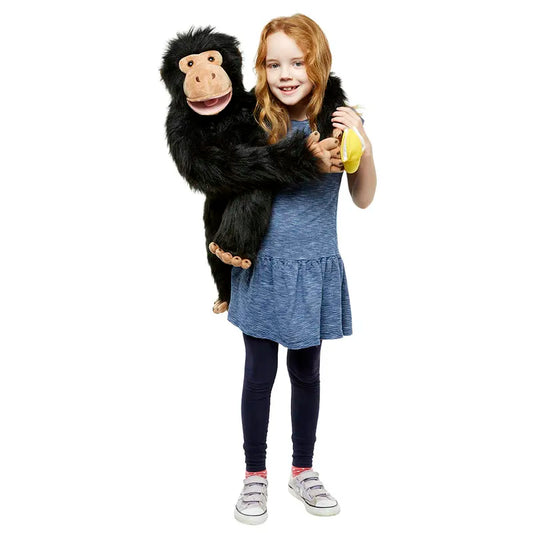 A young girl with long red hair, wearing a blue dress and black leggings, stands against a plain white background, smiling as she holds The Puppet Company Large Puppet Chimp—an expressive monkey puppet that seems ready for some mouth movement. She is also clutching a yellow banana.