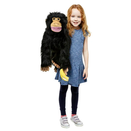 A young girl with red hair, dressed in a blue dress and leggings, is holding The Puppet Company Medium Puppet Primate Chimp. The puppet has black fur and displays a friendly smile, showcasing mouth movement as it holds a yellow banana against the white background.