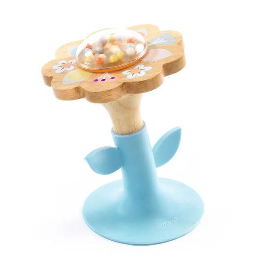 The Djeco Early Years BabySwipi is a flower-shaped wooden rattle with vibrant painted petals and a clear dome holding small beads. It features a blue stem with two leaves and stands upright on a blue base, which includes a suction cup for stability.