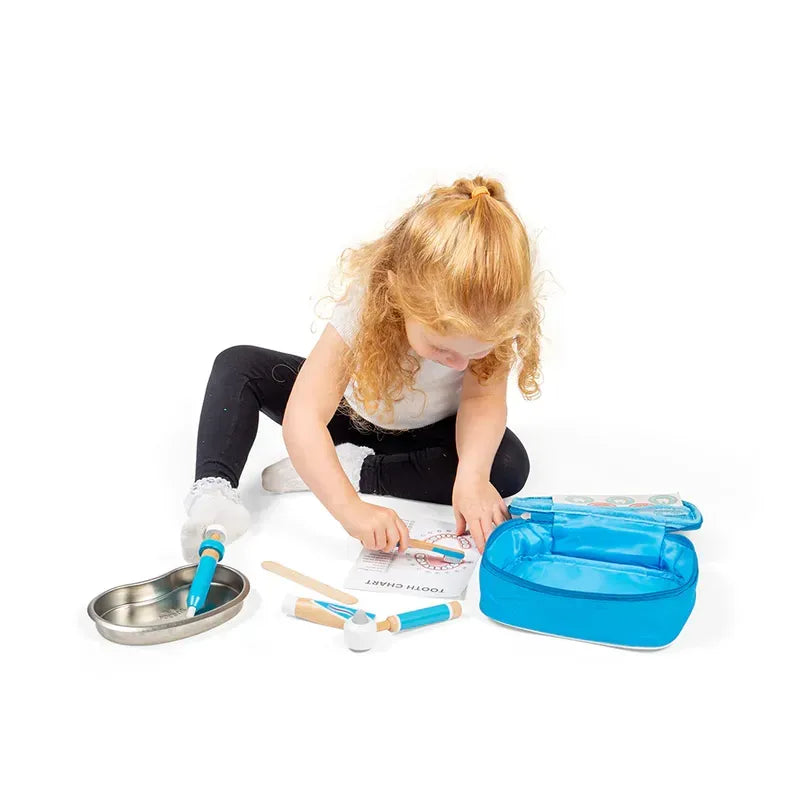 A young, curly-haired child sits on a white floor with the Bigjigs Dentist Kit, playing with various toy dental instruments and examining a "Doctor Patient" sheet.