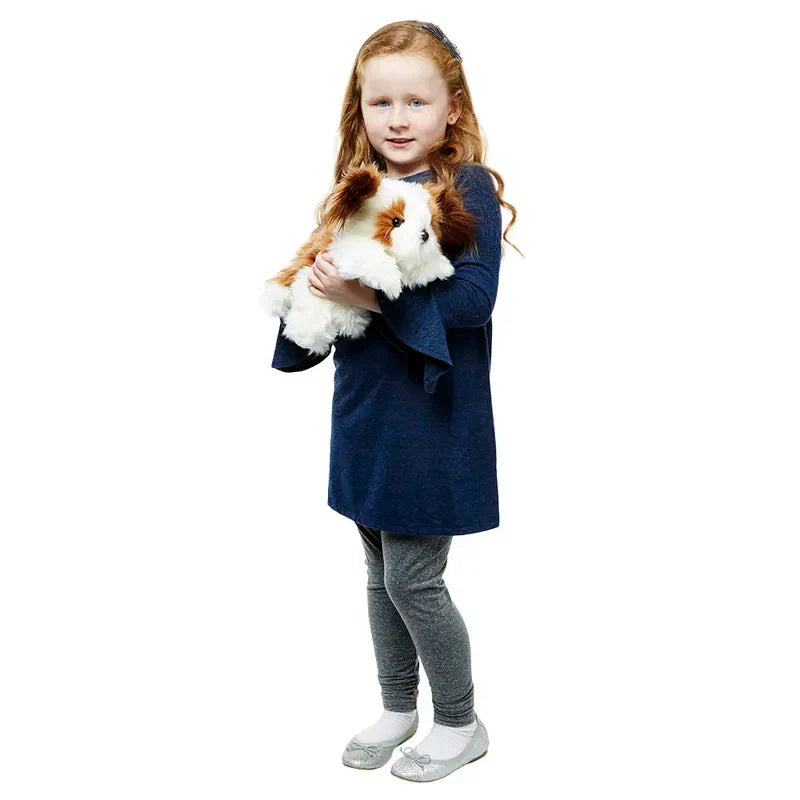 A young girl with long red hair, in a dark blue dress and gray leggings, stands and smiles while holding The Puppet Company Full-bodied Hand Puppet Dog. The background is white.