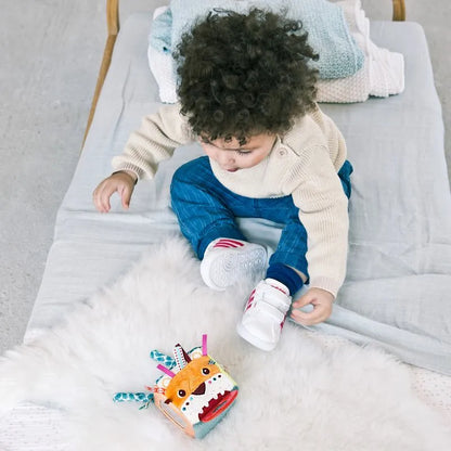 A toddler with curly hair in a beige sweater sits on a bed, captivated by their Lilliputiens I'm Discovering Jack Activity Cube, resting on a white fluffy rug. Wearing blue pants and white sneakers, the child is surrounded by a light blanket and pillows.