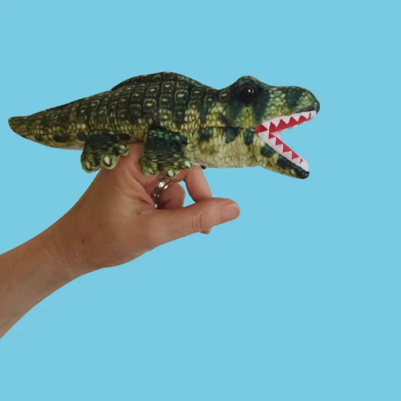 A hand holds The Puppet Company Alligator Finger Puppet from the Little Creatures range, featuring a green open mouth with white teeth, set against a solid light blue background.