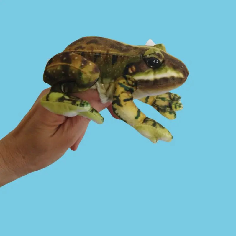 A hand holds The Puppet Company Green Frog Finger Puppet from the Little Creatures Range—a soft, realistic green and brown toy—against a solid light blue background, perfect for storytime fun.