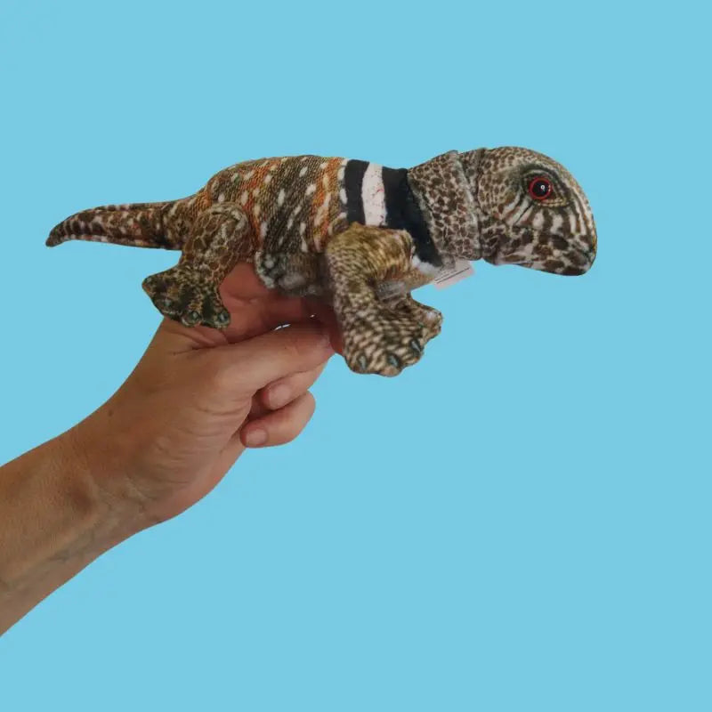 A hand displays The Puppet Company Lizard Finger Puppet – Little Creatures Range, a soft toy with orange, brown, black, and white markings, against a plain light blue background—ideal for nature-loving kids.