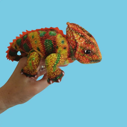 A hand holds The Puppet Company Orange Chameleon Finger Puppet from the Little Creatures Range, displaying vibrant yellow, orange, green, and red patterns against a plain light blue background.