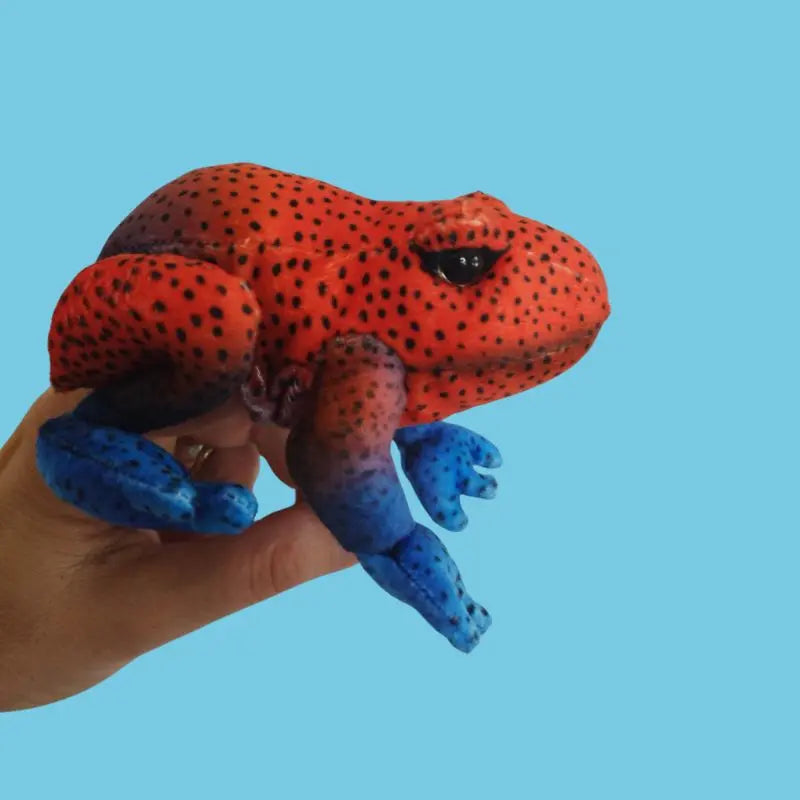 A hand holds The Puppet Company Red Frog Finger Puppet from the Little Creatures Range, showing its vibrant red body with black spots and blue legs against a plain light blue background.