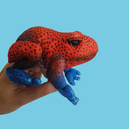 A hand holds The Puppet Company Red Frog Finger Puppet from the Little Creatures Range, showing its vibrant red body with black spots and blue legs against a plain light blue background.