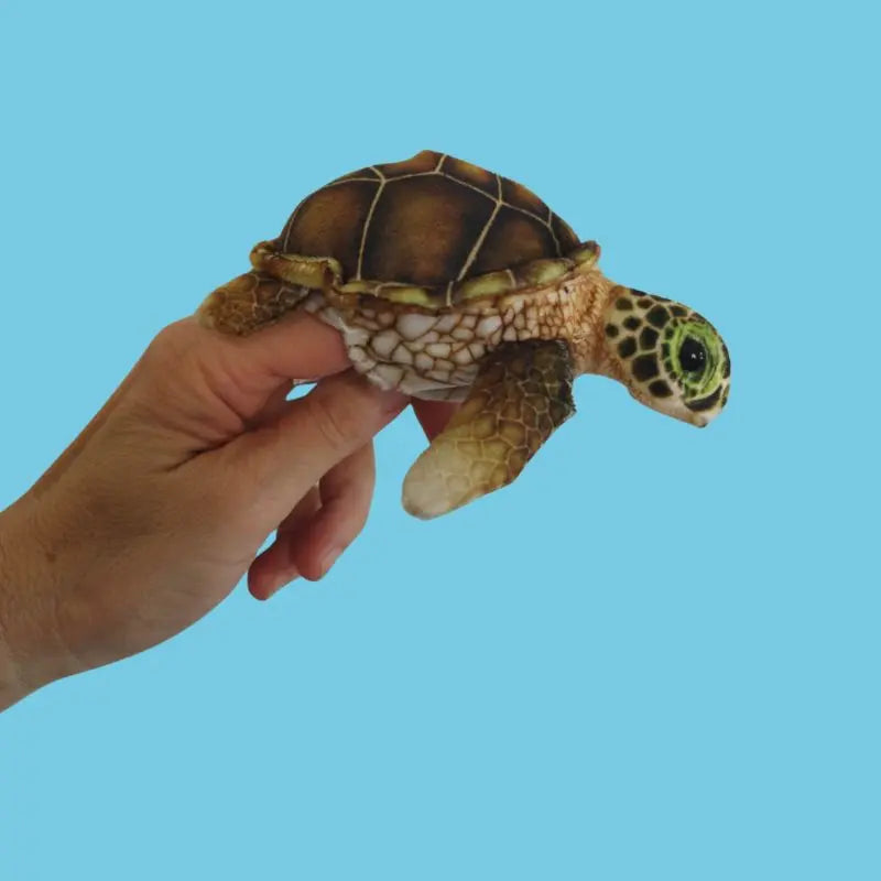 A hand holds The Puppet Company Turtle Finger Puppet from the Little Creatures Range against a plain light blue background, inviting quiet play and imaginative adventures.