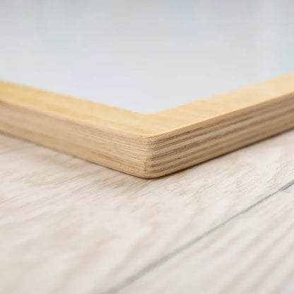 Close-up of the corner of the Bigjigs Magnetic Board on a wooden surface, ideal for showcasing creativity. Its light wood finish with visible grain contrasts sharply with the slightly textured surface beneath.
