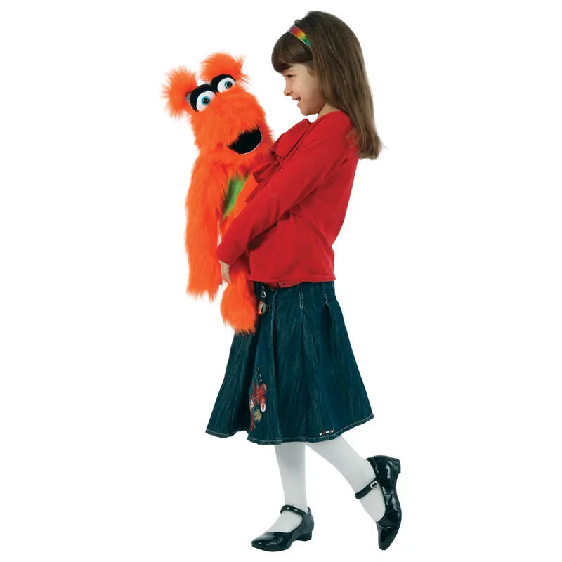 A young girl in a red sweater and denim skirt smiles while holding The Puppet Company Orange Monster puppet, featuring googly eyes and a big mouth, against a white background.