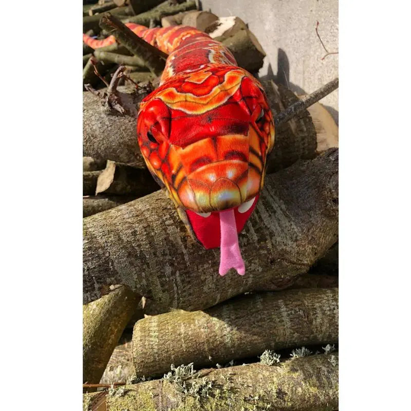 The Puppet Company Large Creature Snake Orange is a realistic, brightly colored orange and red snake hand puppet with its tongue out, resting on a pile of logs.