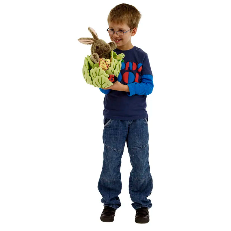 A young boy with glasses, in a blue shirt and jeans, holds The Puppet Company Hide-Away Rabbit— a soft cuddly rabbit in a green basket with foliage and ladybugs. He smiles at the plush toy against a plain white background.
