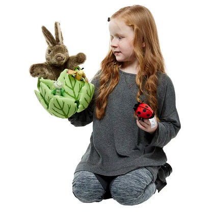 A young girl with long red hair kneels on the floor, delighting in her finger puppets. She holds a frog puppet and a ladybug toy, while The Puppet Company Hide-Away Rabbit is perched on her other hand. Her gray sweatshirt and patterned leggings add to her charm as she smiles at these imagination toys.