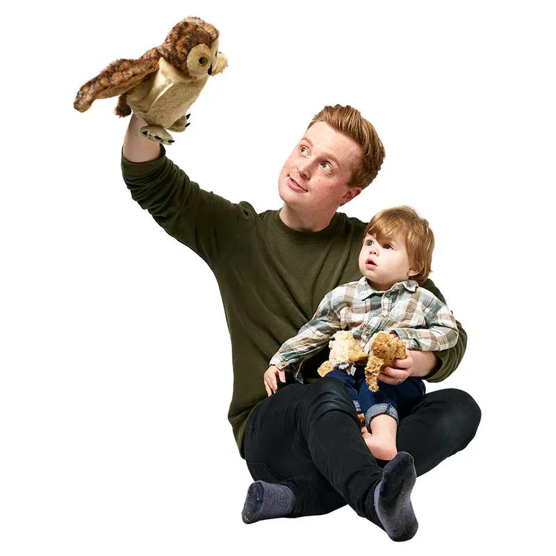 A man sits on the floor with a toddler on his lap, engaging in imaginative play using The Puppet Company Hide-Away Tawny Owl puppet. The toddler, holding a small toy, curiously watches the puppet. Both are casually dressed against a plain white background.
