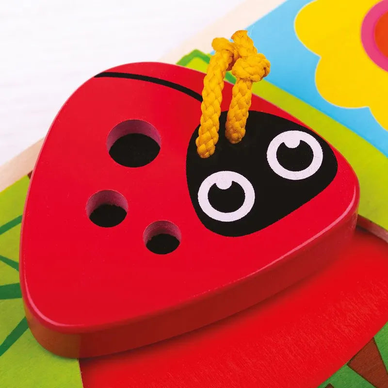A close-up of a colorful wooden puzzle piece from the Touch and Feel Bugs Puzzle shaped like a red ladybug features three black holes and a yellow string through one hole. The ladybug has a cute face with big eyes against a textured background with bright colors and floral shapes.