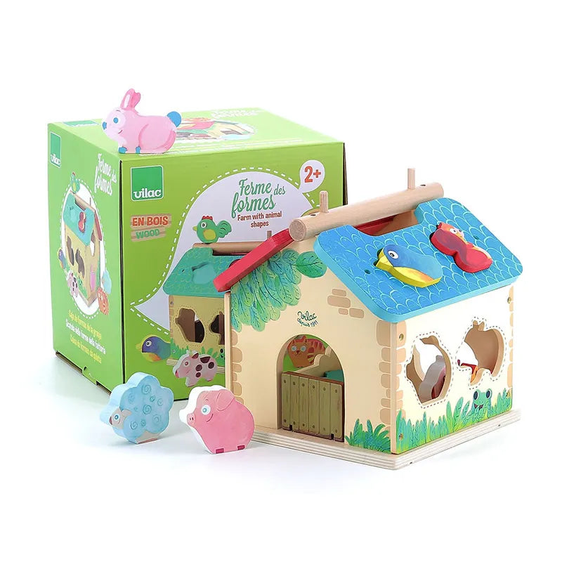 The Vilac Farm With Shapes wooden toy farmhouse, featuring vibrant animal shapes, comes with a green and yellow box that showcases an image of the farmhouse and specifies it's suitable for ages 2 years and up. A pink rabbit piece is perched on the box, while blue and pink animal pieces are scattered on the table.