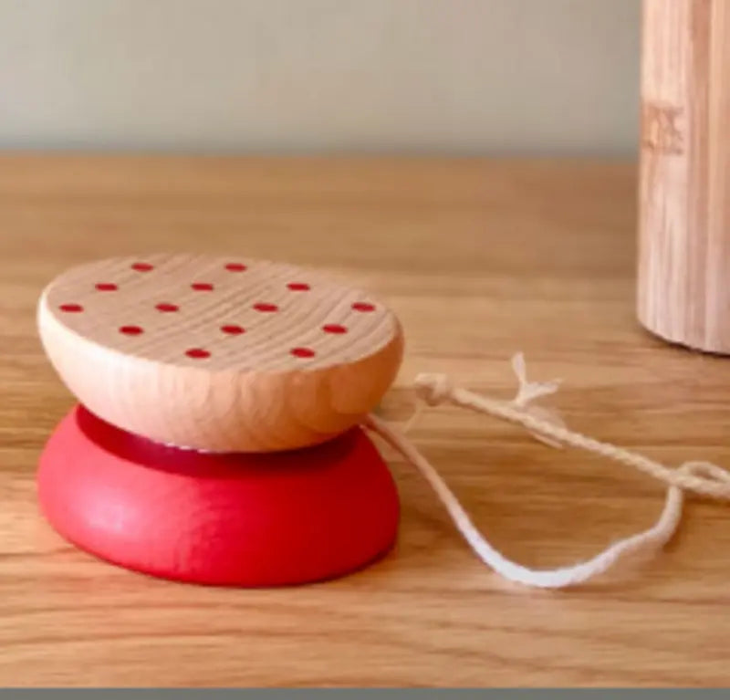 The Wooden Yo-Yo Red, made from smooth beechwood with a red base, natural top, and playful red dots, sits on a wooden surface. Its white string offers endless screen-free fun.